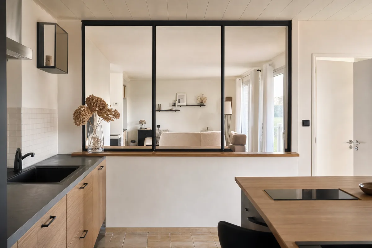 Verrière d&rsquo;atelier en acier noir posée sur un muret bas entre une cuisine claire et un séjour, lumière traversante