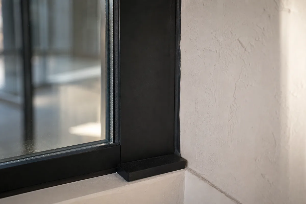 Détail d&rsquo;un montant de verrière en acier noir raccordé à un mur en enduit clair, joint de finition et vitrage feuilleté